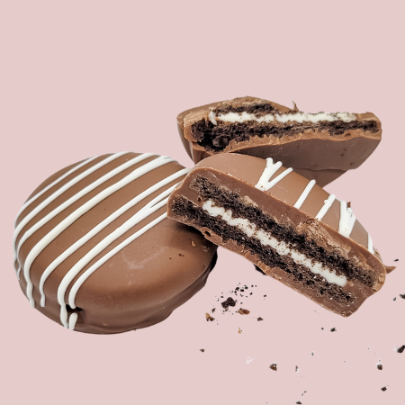 Four chocolate dipped Oreos with white drizzle on a pink background, one broken to show cream filling.