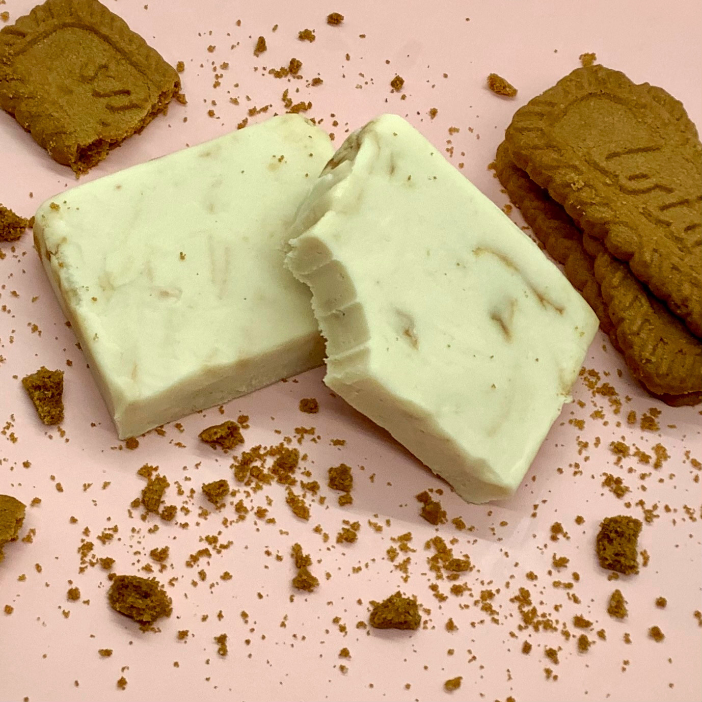 Pale green Biscoff cheesecake fudge block broken in half on a pink surface with cookie crumbs nearby