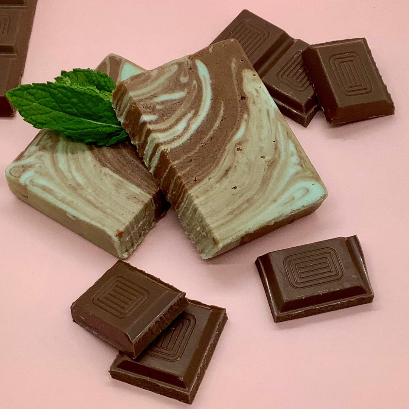 Swirled chocolate mint fudge blocks with mint leaves and pieces of dark chocolate on a pink background.