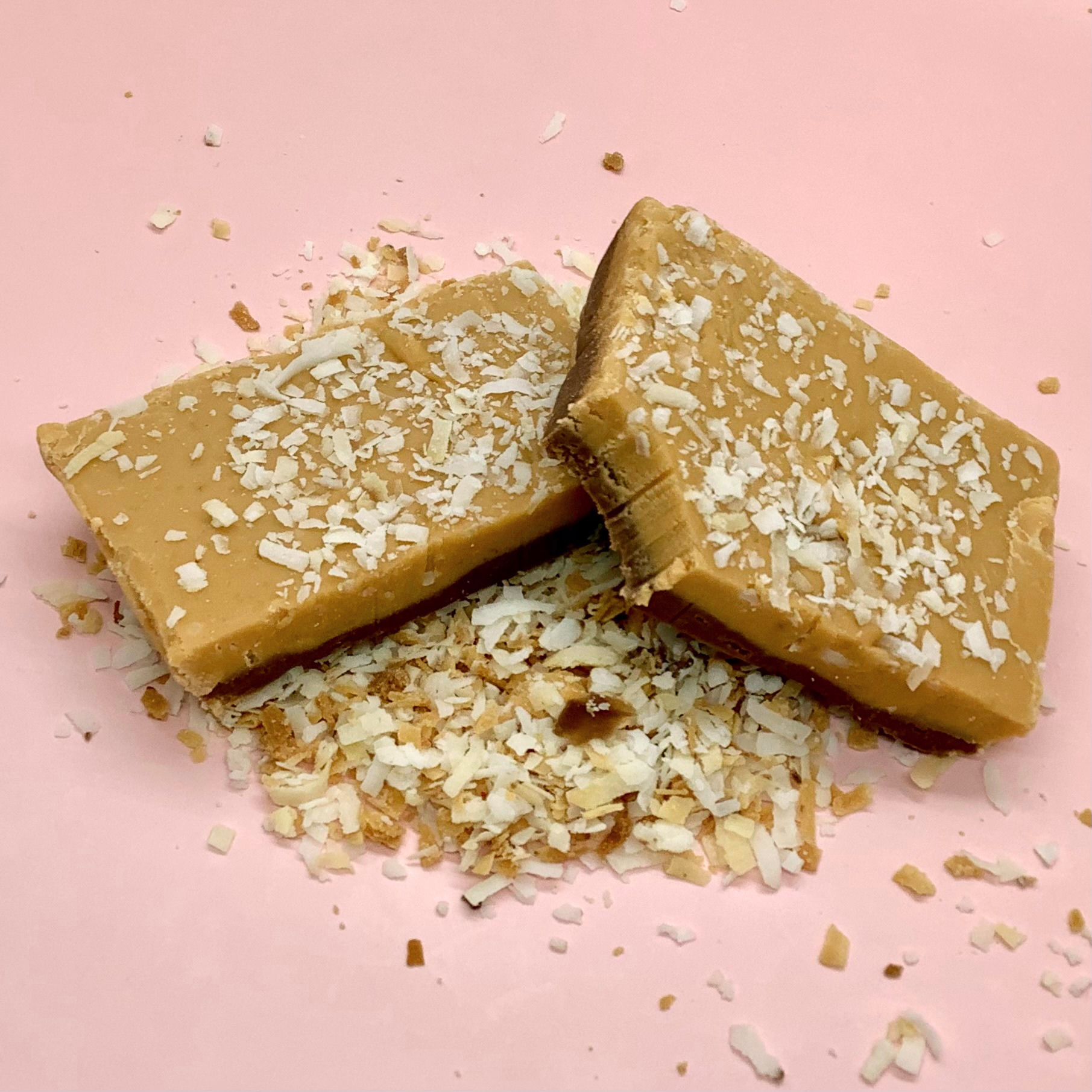 Two coconut caramel fudge bars dusted with coconut flakes on a pink background