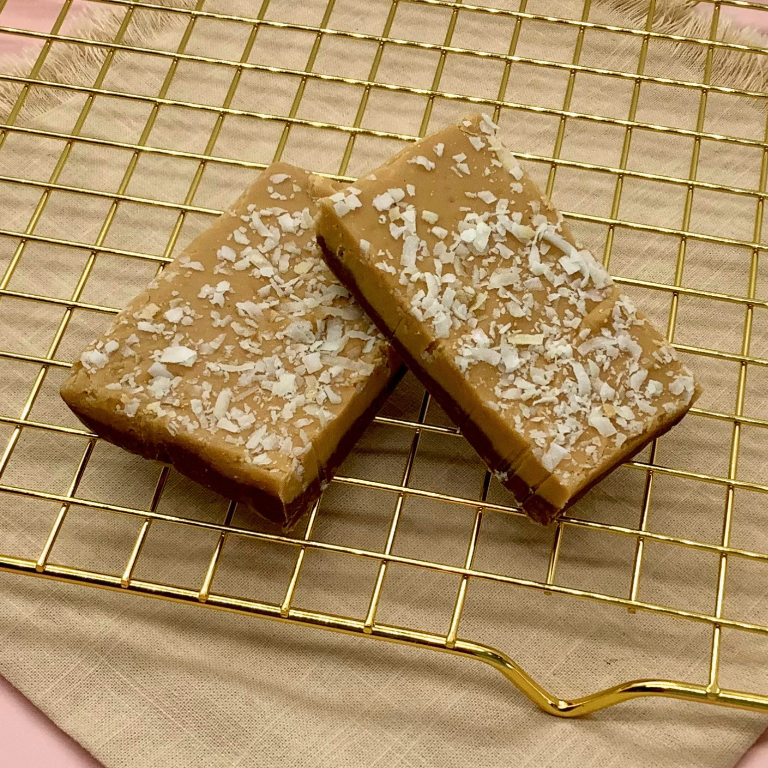 Two coconut caramel fudge squares topped with coconut flakes on a gold wire cooling rack.