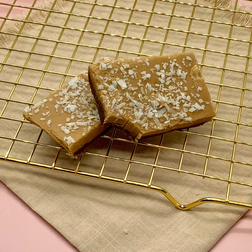 Two coconut caramel fudge squares topped with coconut flakes on a gold cooling rack.