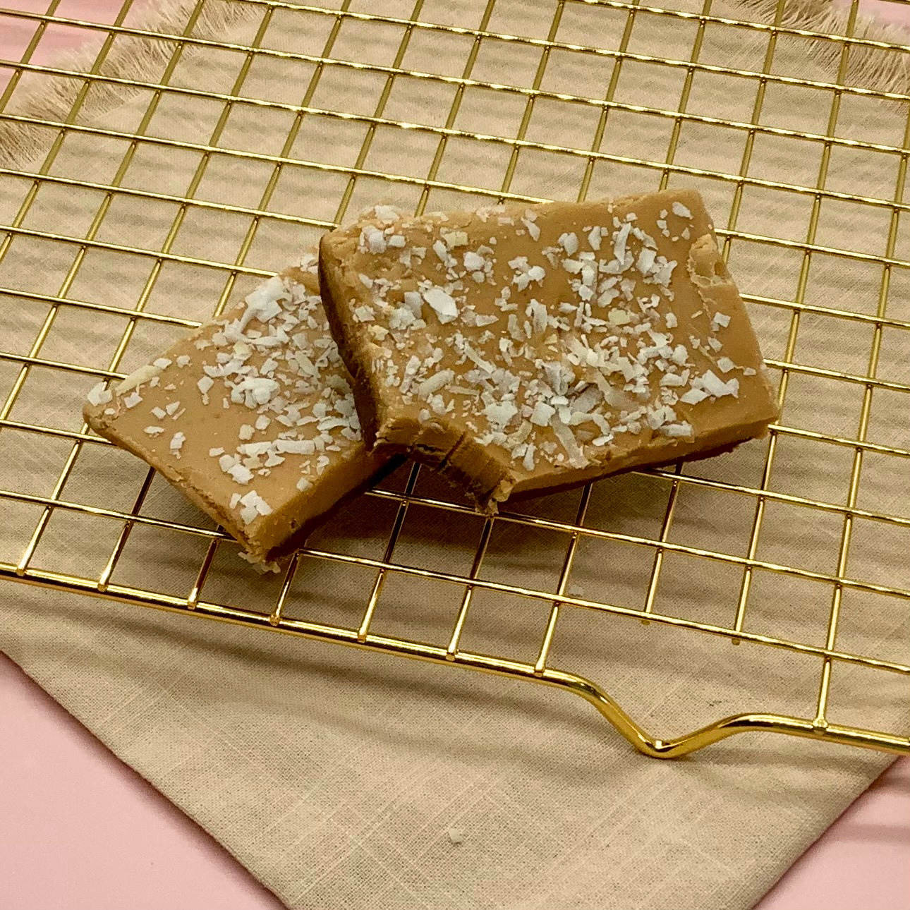 Two coconut caramel fudge squares topped with coconut flakes on a gold cooling rack.