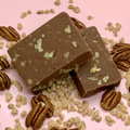 Two chocolate toffee fudge squares with pecans on a pink background and scattered pecan pieces